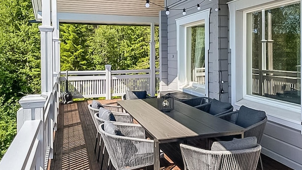 Een overdekt terras met een lange tafel en stoelen, omgeven door een witte balustrade, met uitzicht op een bebost gebied.