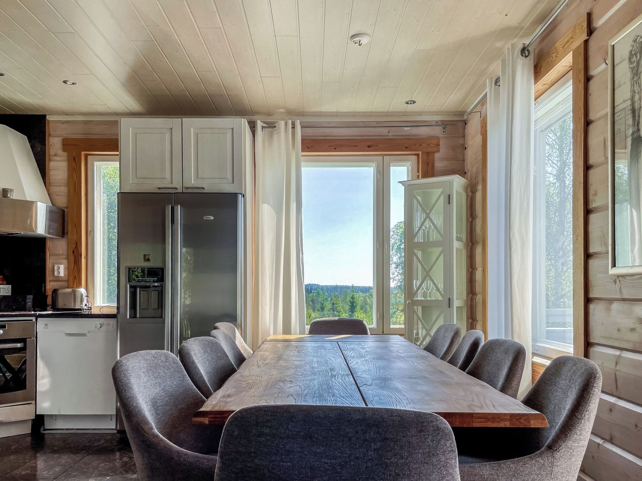 Une cuisine moderne dotée d’un réfrigérateur en acier inoxydable, de meubles blancs et d’un coin repas avec une table en bois et des chaises à revêtement gris.