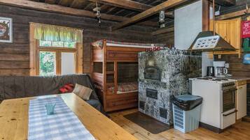 A rustic cabin interior with a wooden dining table, a bunk bed, a stone fireplace, and a kitchenette.