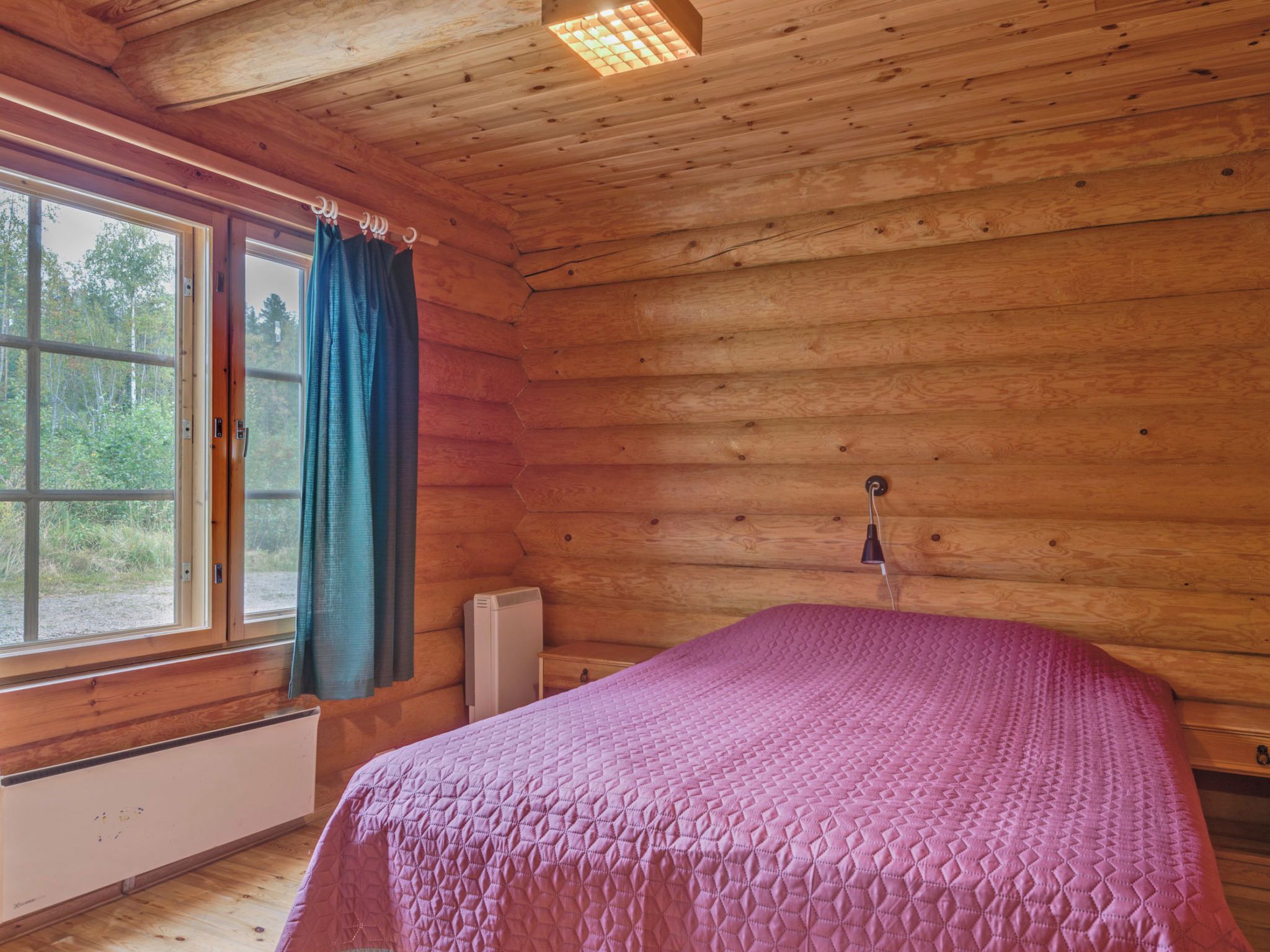 Ein Schlafzimmer einer Holzhütte mit einem Bett, einem Fenster mit Vorhängen und einer Heizung.