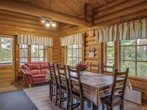 Interior de una cabaña de madera con una mesa de comedor, sillas, un sofá de cuero y grandes ventanales con vistas al bosque.