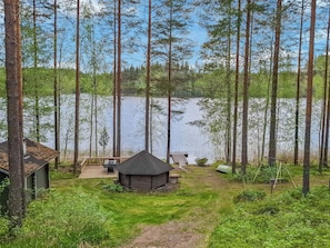 A cabin with a deck and a small shed, surrounded by tall trees and a lake.