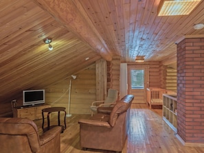 A log cabin interior with a loft bedroom, a television, leather furniture, and a brick fireplace.
