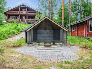 A wooden cabin with a sloped roof and a stone foundation, surrounded by a forested area.
