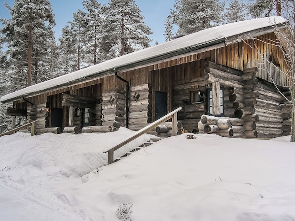 A log cabin with a snow-covered roof, wooden stairs leading to the entrance, and a clear sky in the background.
