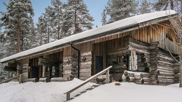 Lumihunnun peittämä hirsimökki, jonka ulkoseinät ovat puuta, ja joka on lumisten puiden ympäröimä.