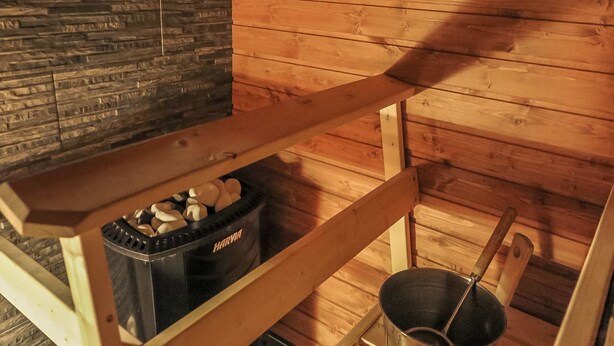 A sauna room with wooden walls, a stone wall, and a wooden bench with a black container holding stones. - Soidinaukea b 20 by Interhome (Inari)