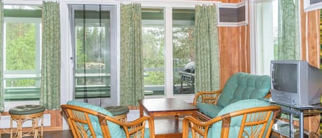 Ein Zimmer mit Holzwänden, Korbmöbeln, einem Fernseher und großen, mit Vorhängen versehenen Fenstern.