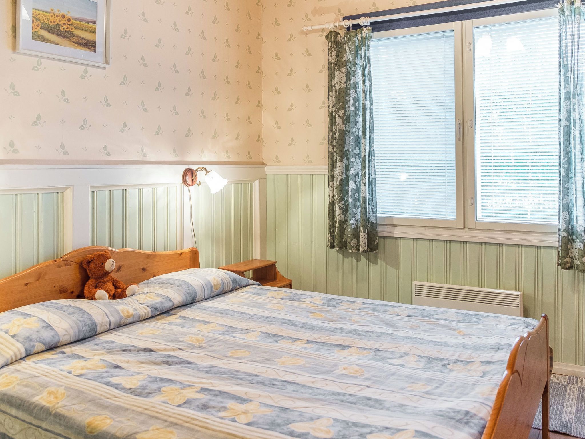 A bedroom with a bed, a nightstand, a teddy bear, a wall-mounted lamp, and a window with curtains.