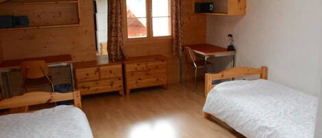 A wooden cabin room with two beds, a desk, a chair, a dresser, and a window with curtains.