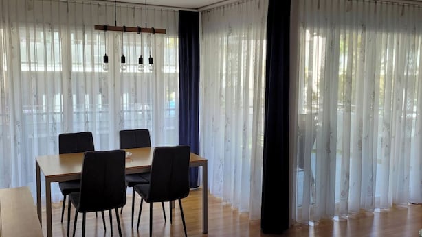 Ein Essbereich mit Holztisch und -stühlen, großen Fenstern mit transparenten Vorhängen und einem dunklen Vorhang an einer Seite.
