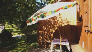 En træterrasse med et bord og bænk under en farverig parasol, omgivet af træer og et træhegn.