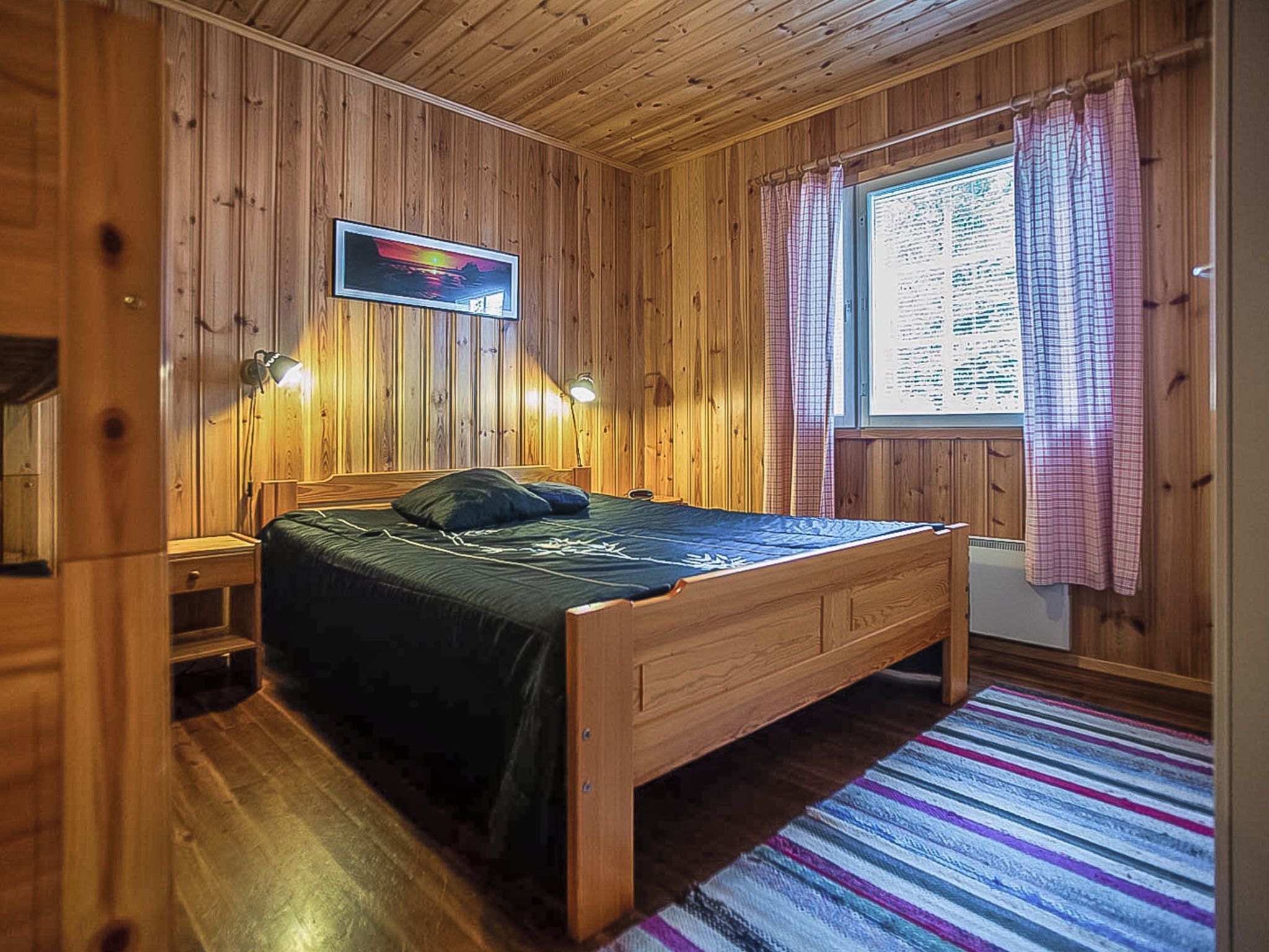 A wooden-paneled room with a bed, two bedside lamps, a window with curtains, and a wall-mounted picture.