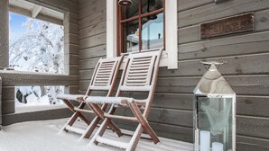 A wooden porch with two wooden chairs covered in snow, a glass lantern, and a window with a view of snow-covered trees.