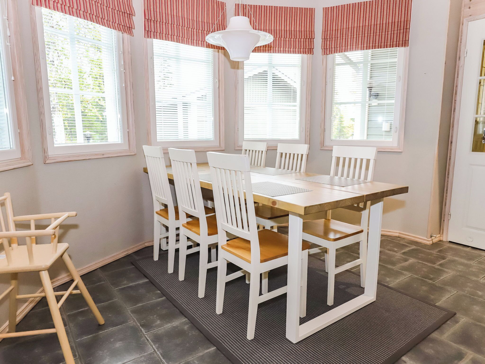 A dining area with a wooden table and white chairs, large windows with blinds, and a white pendant light.