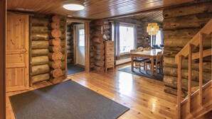 Interior de una cabaña de madera con una mesa de comedor, sillas, una escalera y una alfombra grande en el suelo.