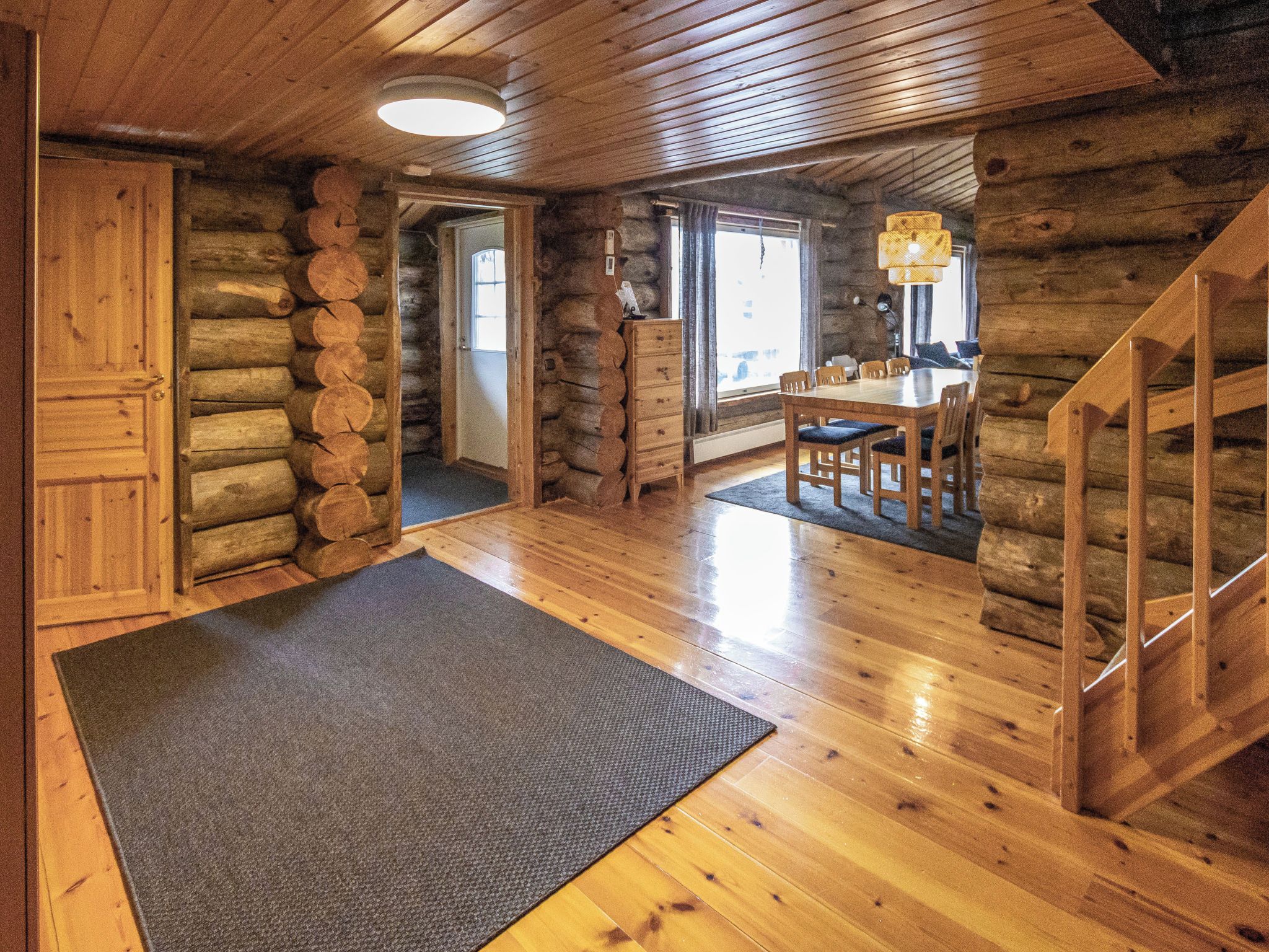 A log cabin interior with a wooden dining table, chairs, a staircase, and a large rug on the floor.