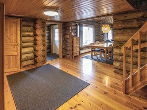 A log cabin interior with a wooden dining table, chairs, a staircase, and a large rug on the floor.