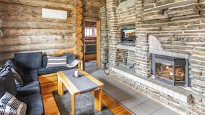 Interior de una cabaña de troncos con chimenea de piedra, un sofá y una mesa de madera.