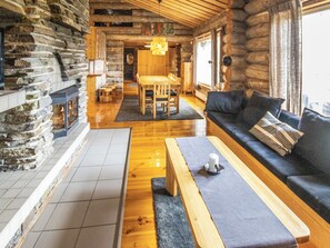 Het interieur van een blokhut met een stenen openhaard, een houten eettafel en een bank met kussens.