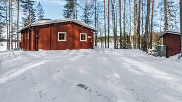 Una cabaña de madera con un ventanal y una puerta, rodeada de nieve y árboles altos.