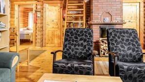 A cozy log cabin interior with a fireplace, two patterned armchairs, a bookshelf, and a staircase leading to a loft.