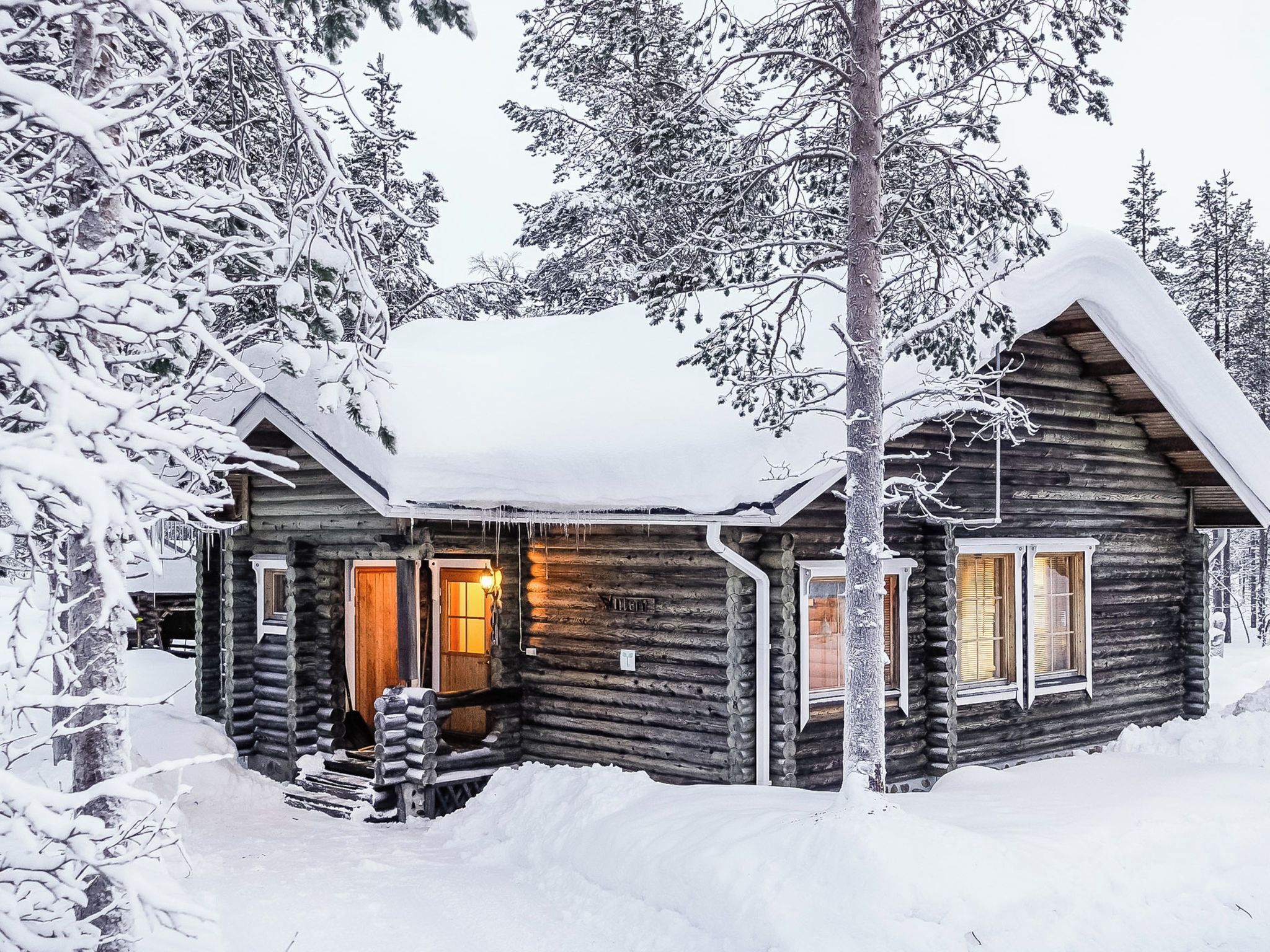 Une cabane en rondins avec un porche en bois, un intérieur éclairé et un environnement enneigé.