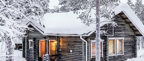 A log cabin with a wooden porch, a lit interior, and snow-covered surroundings.