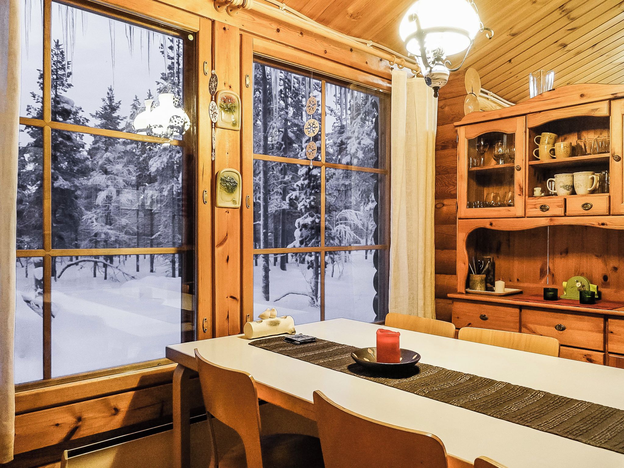 L'intérieur d'un chalet en bois comprend une table à manger, des chaises, une lampe et offre une vue sur une forêt enneigée grâce à de grandes fenêtres.