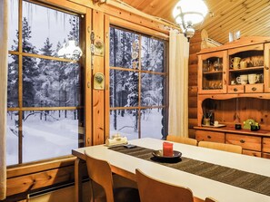 A wooden cabin interior with a dining table, chairs, a lamp, and a view of a snowy forest through large windows.