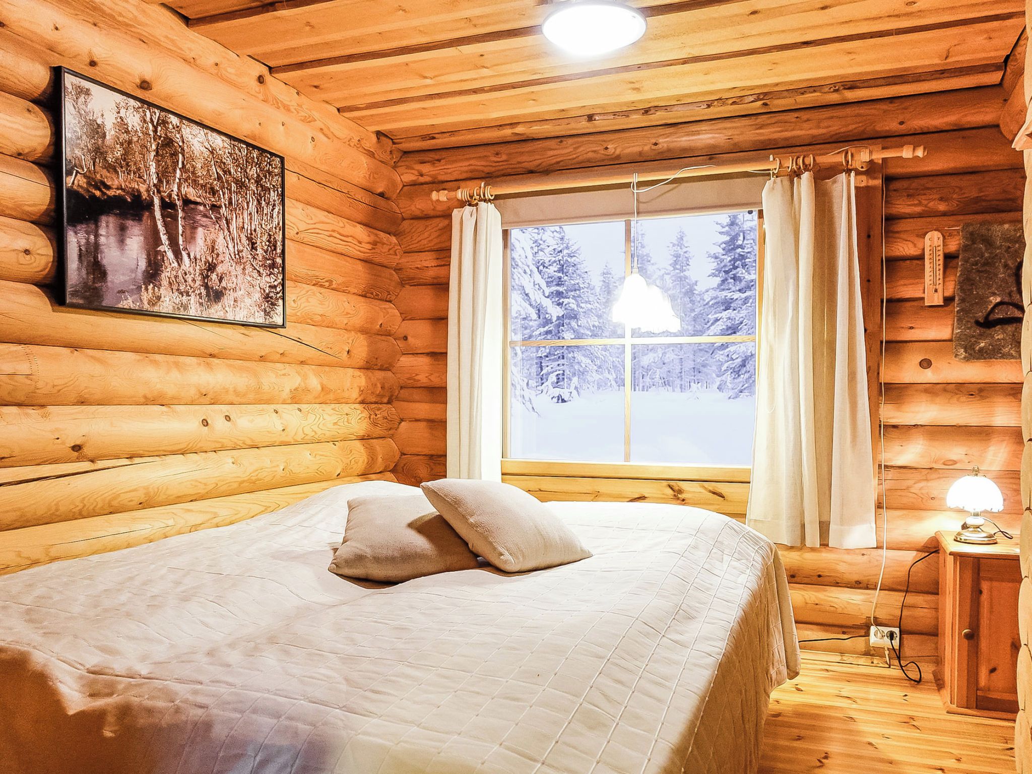 Une chambre dans une cabane en rondins, avec un lit, une fenêtre avec des rideaux, une lampe suspendue et un tableau encadré au mur.