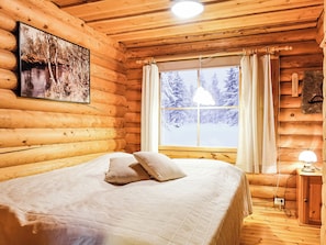 Un dormitorio tipo cabaña de madera con cama, ventana con cortinas, lámpara colgante y un cuadro en la pared.