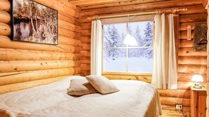 A log cabin bedroom with a bed, a window with curtains, a hanging light fixture, and a painting on the wall.