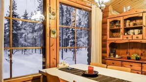 A cozy cabin interior with a dining table, chairs, wooden cabinets, and a view of snow-covered trees through large windows.