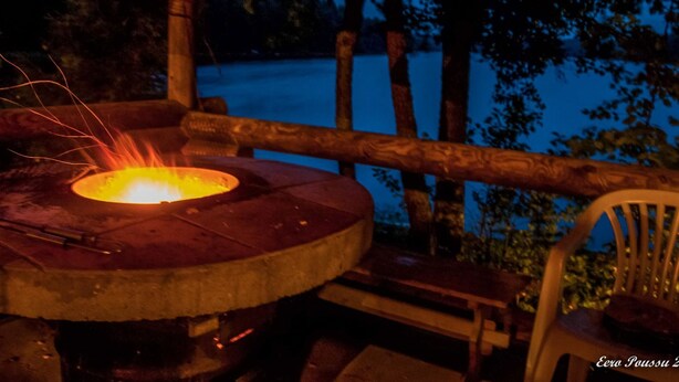 A wooden outdoor seating area with a round table and a fire pit. - Kalliohonka by Interhome (Hämeenlinna)