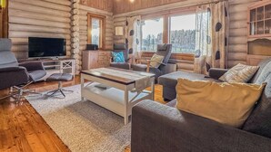 A log cabin interior with a TV, sofa, armchair, and a view of the forest through large windows. - Kalliohonka by Interhome (Hämeenlinna)