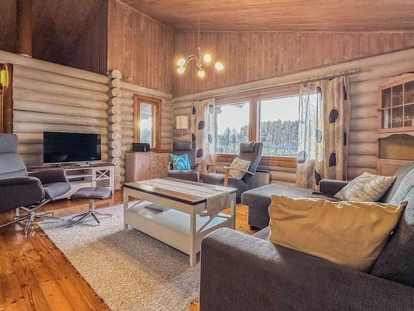 Ein Holzblockhaus-Interieur mit Fernseher, Sofa, Sessel und Blick auf den Wald durch große Fenster.