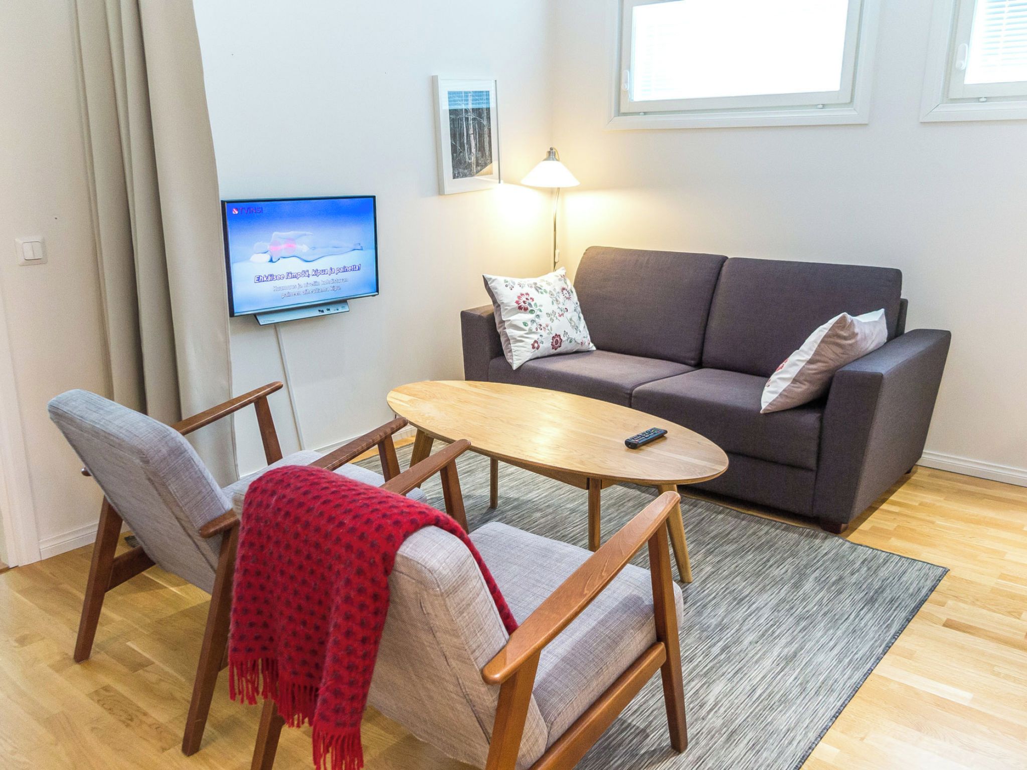 Een woonkamer met een grijze bank, twee fauteuils, een houten salontafel, een aan de muur bevestigde televisie en een vloerlamp.
