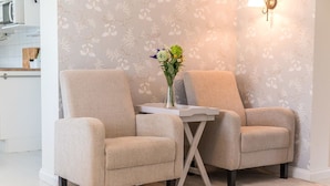 To beige lænestole med et lille bord imellem, en vase med blomster, og en vægmonteret lampe.