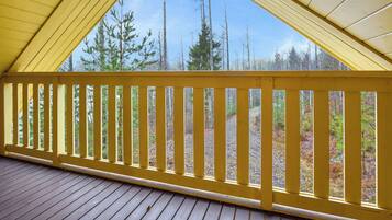 Una terraza de madera con barandal amarillo, que conduce a un espacio cubierto con vista a árboles y cielo.