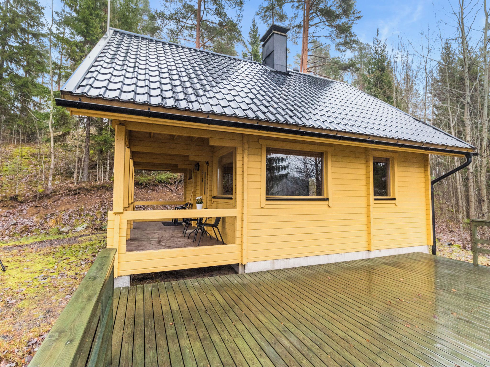 En gul stuga med en inglasad veranda och ett trädäck, omgiven av träd.