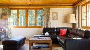 Ein gemütliches Wohnzimmer mit Holzbalkendecke, einem schwarzen Ledersofa, einem Holztisch und einem großen Fenster mit Vorhängen.