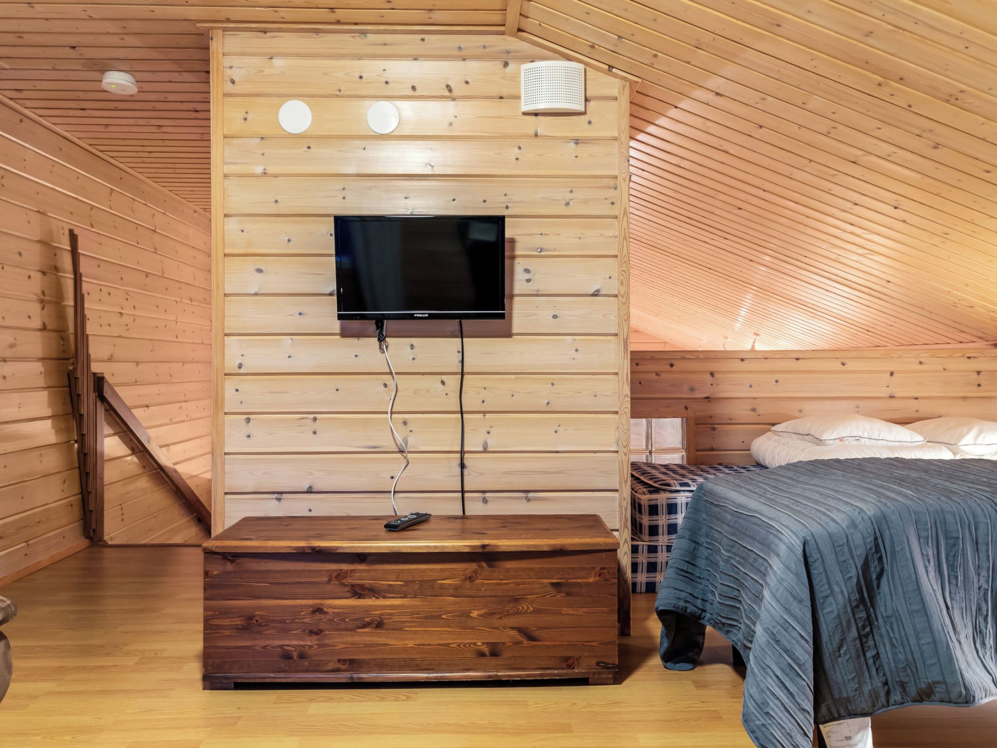 A room with a television mounted on a wooden wall, a bed with a blue blanket, and a wooden bench.