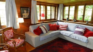 Een gezellig cabin interieur met een houten plafond, een grijze modulaire bank met rode en witte kussens, een roodgeel patroon tapijt, en een houten stoel met een rood kussen.