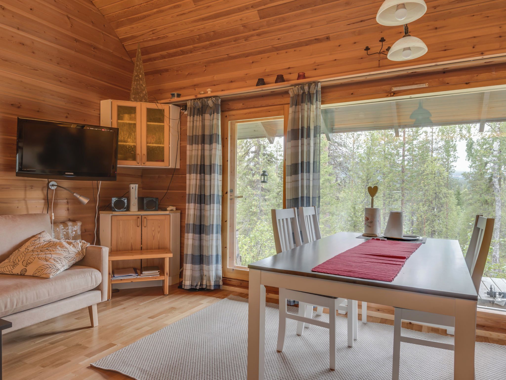 A cozy cabin interior with a wooden ceiling, a television mounted on the wall, a dining table with chairs, and a window offering a forest view.