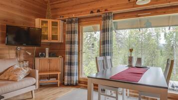 A cozy cabin interior with a wooden ceiling, a television mounted on the wall, a dining table with chairs, and a window offering a forest view.