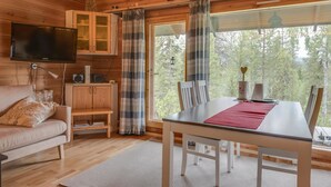L’intérieur chaleureux d’un chalet, avec un décor en bois, une table à manger entourée de chaises, un canapé, un téléviseur fixé au mur et une grande fenêtre donnant sur la forêt.