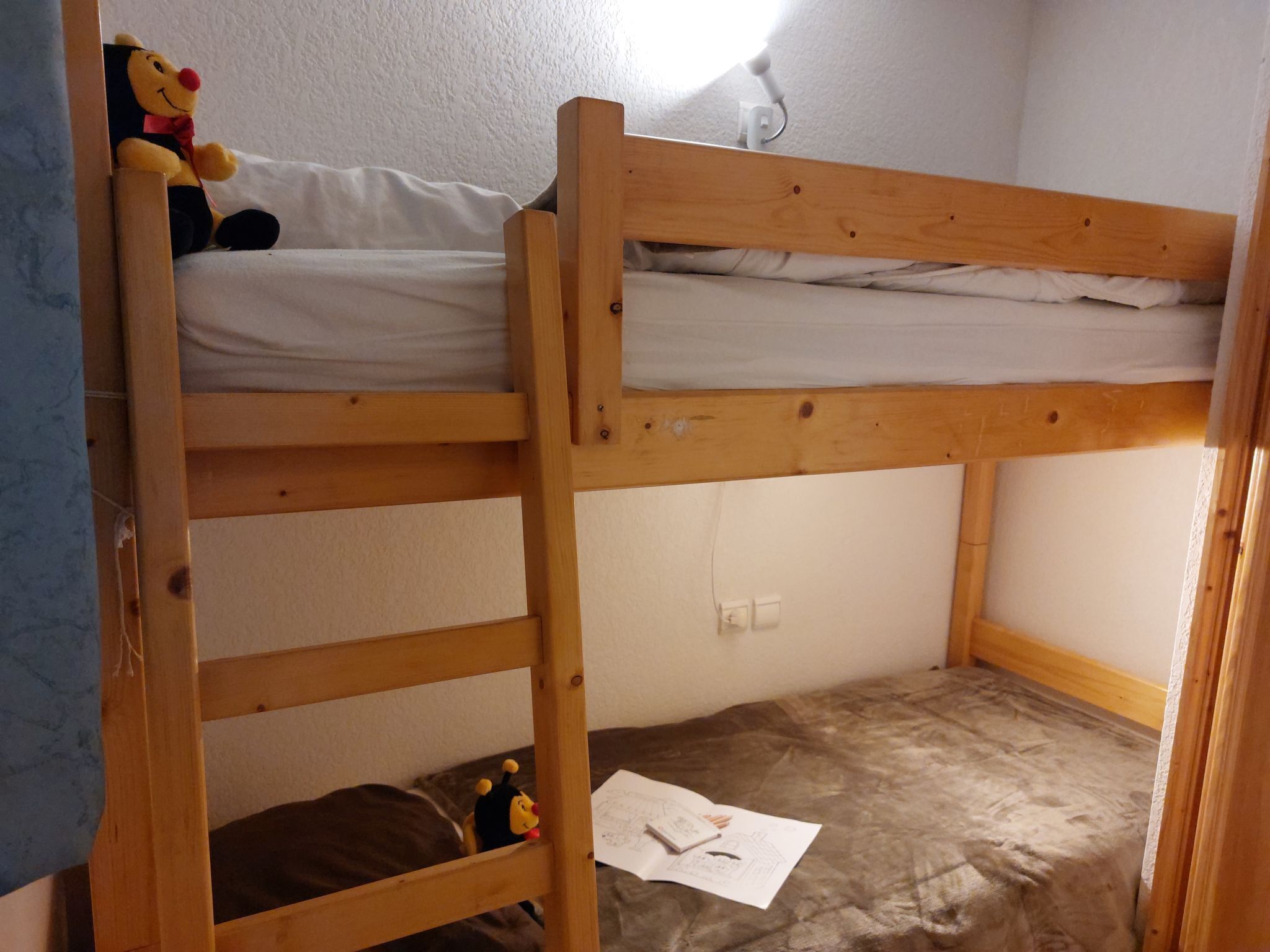 A bunk bed room with a wooden frame, a white mattress on the top bunk, and a brown mattress on the bottom bunk. There is a small toy on the top bunk and a piece of paper on the floor.