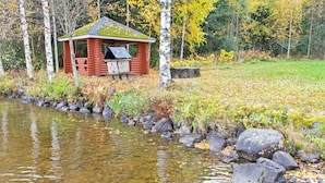 En stuga av trä med en liten veranda vid en klar bäck, omgiven av träd och stenar.