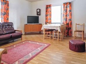 A living room with a sofa, a television, a dining table with chairs, a wooden cabinet, and a rug.